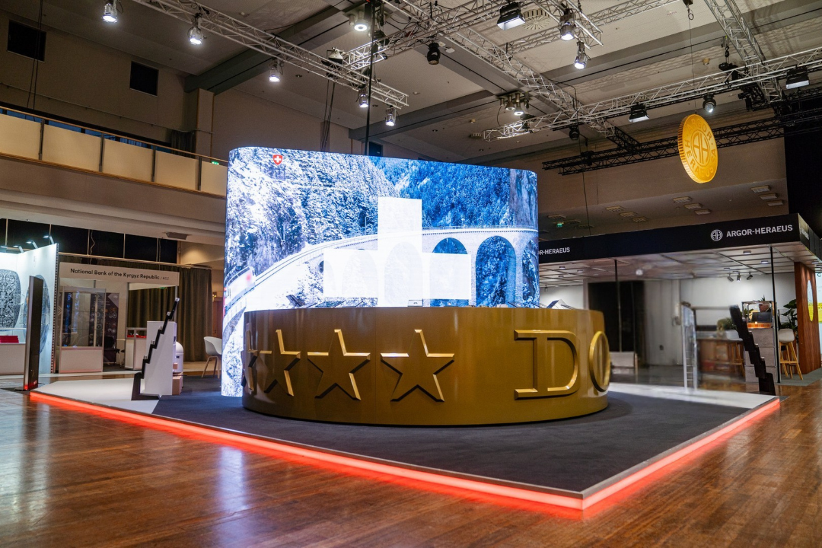 Individueller Messestand von Swissmint mit großer, gebogener LED-Wand und goldener Theke der Eidgenössischen Münzstätte.